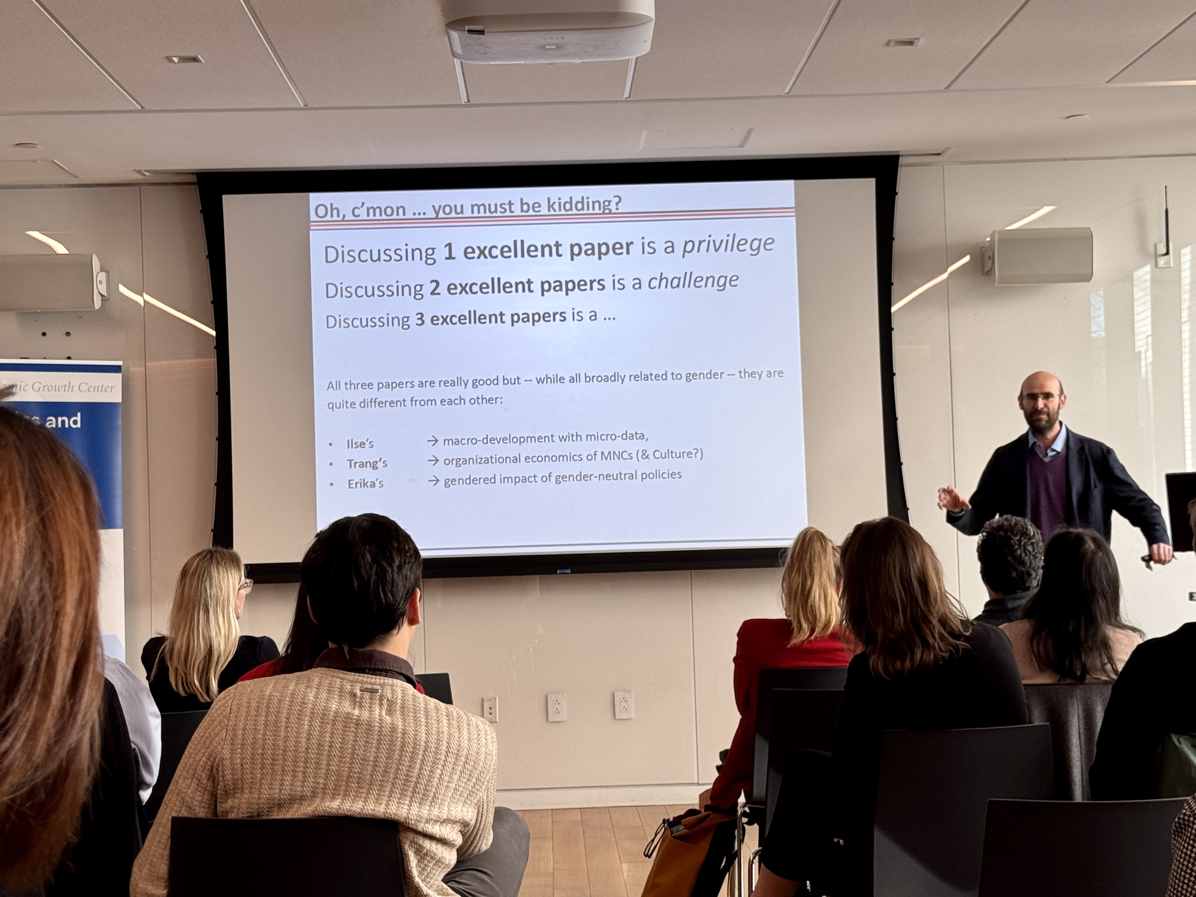 A man in a black jacket presents at the front of a classroom