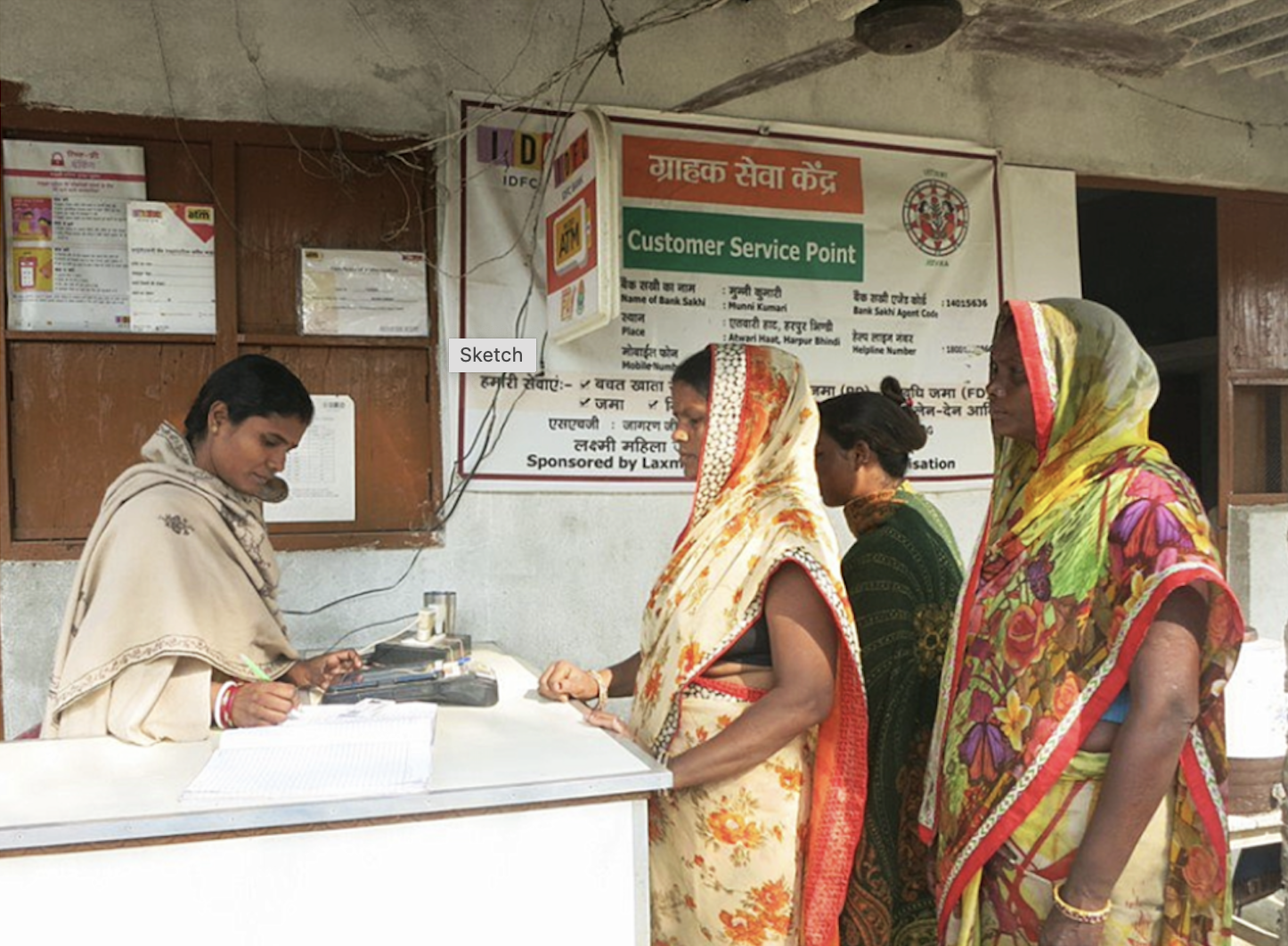 Three Indian women standing in line at a customer service center, while another woman is behind the register helping them. 