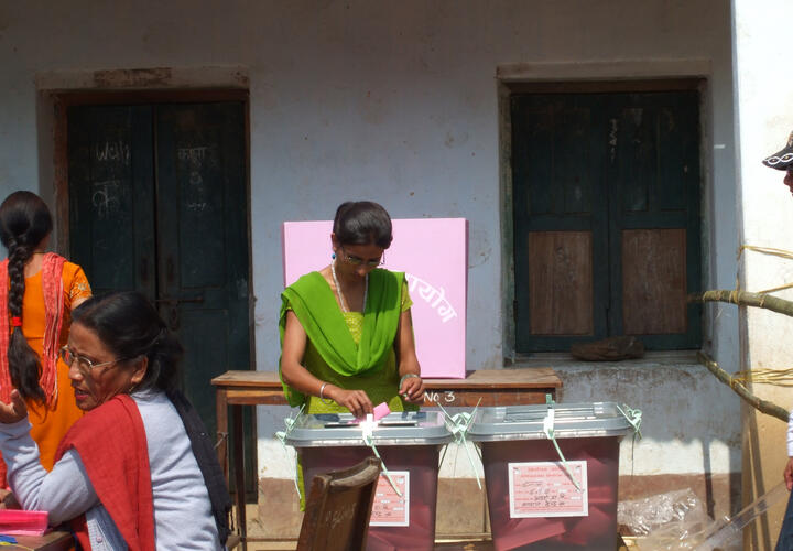 A woman in a green dress places a pink slip of paper into a brown box.