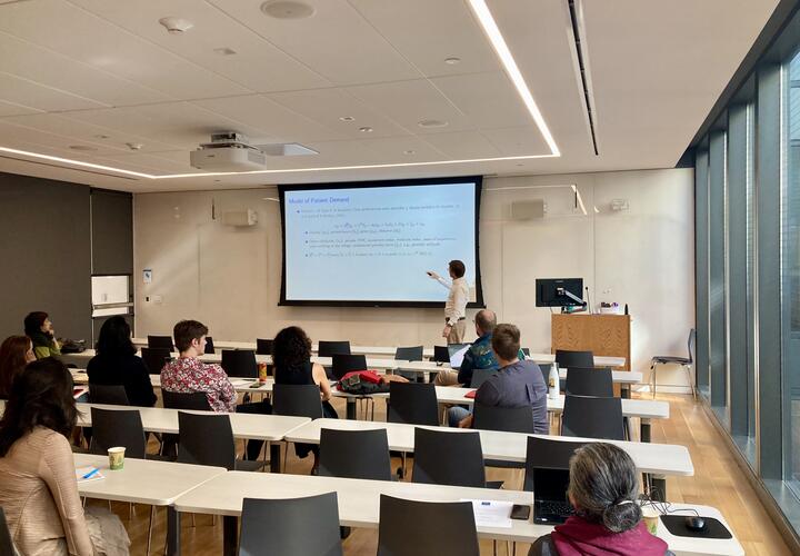 A man in a white shirt points to a slide during a presentation to a classroom of economics researchers