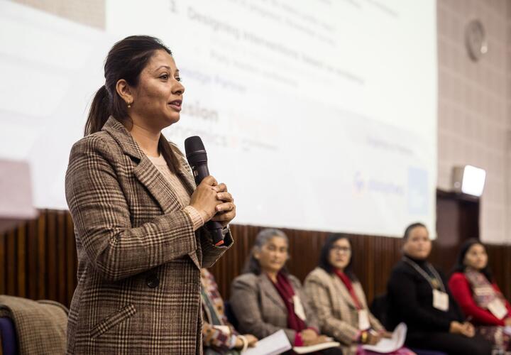 A woman in a grey jacket addresses an audience, with a panel of speakers behind her