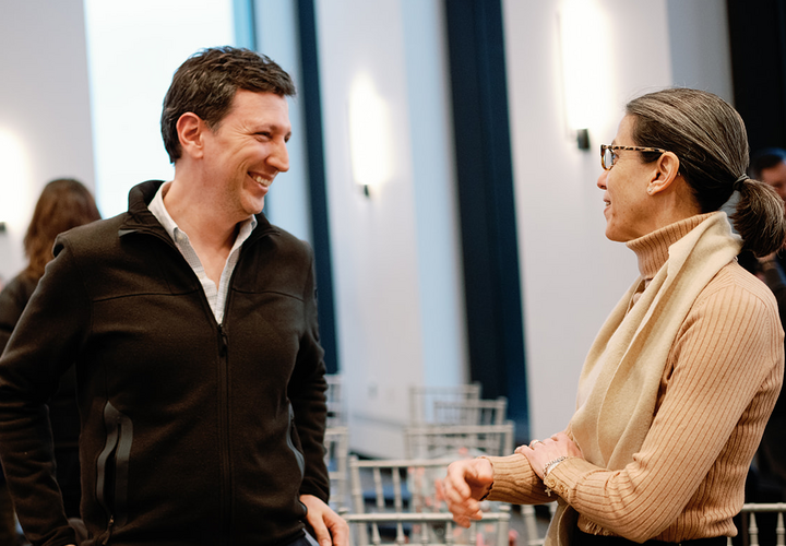 A man in a black jacket and a woman in a tan sweater converse at an event