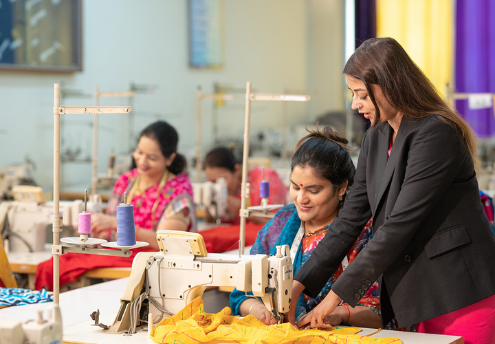 A woman in a black suit observes a woman in a colorful dress at a sewing machine
