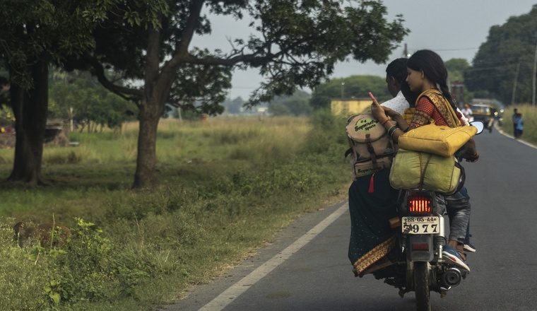 Two people riding on a motorcycle away from the viewer, one holding a cellphone